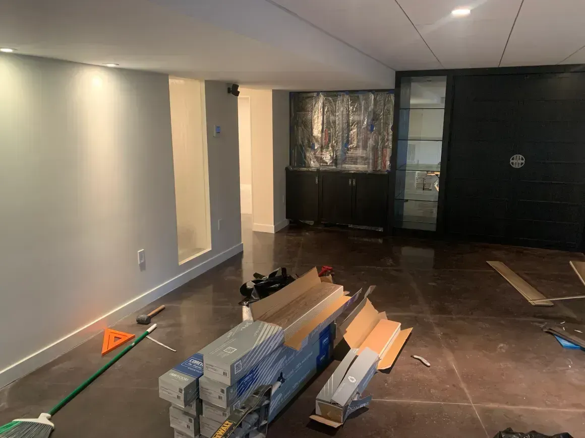 Interior of a room under construction with gray walls, dark floor, and stacked boxes of flooring.