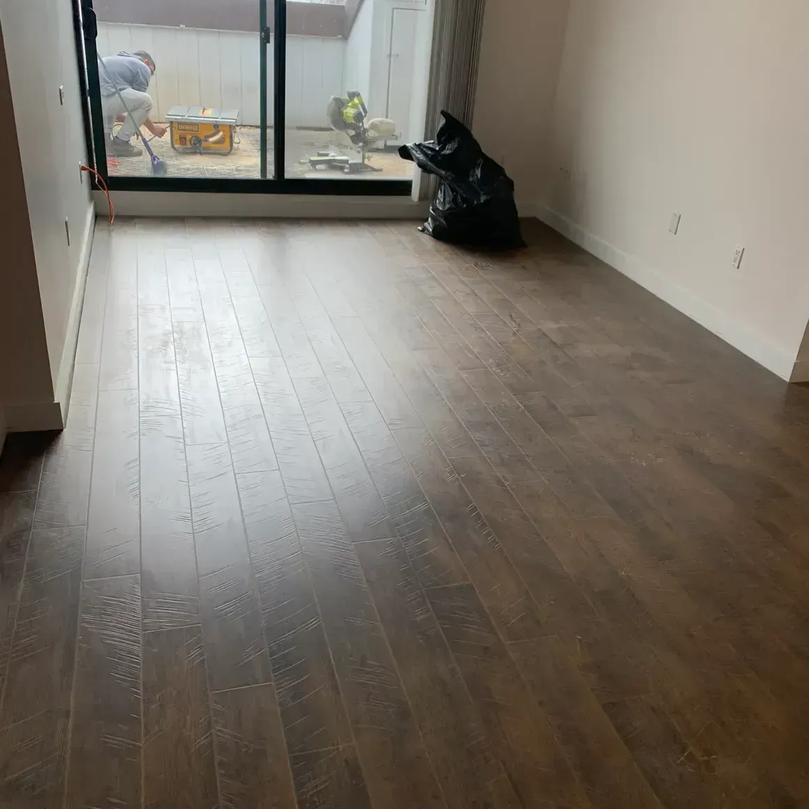 Dark wood floors in an empty room with a sliding glass door open to an outdoor area; a black trash bag sits in the room.