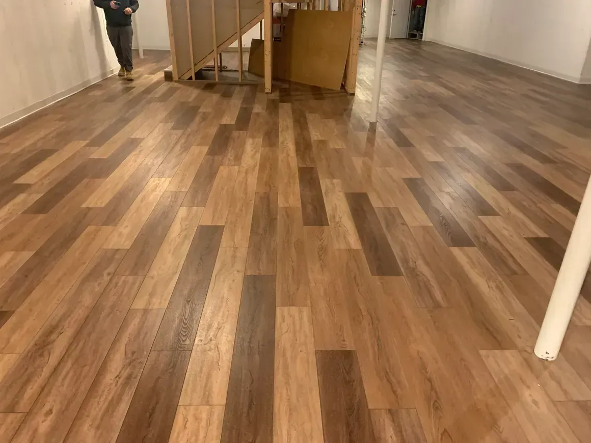 Newly installed wood-look flooring in a room with a staircase and white walls. Two people are present in the background.