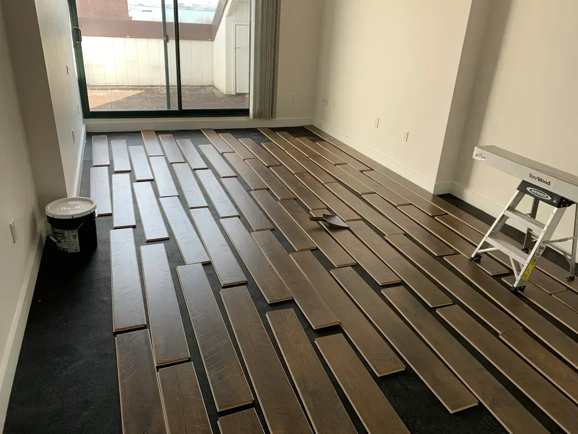 Wooden floor planks laid out on a dark underlayment in a room with a window and a paint can.