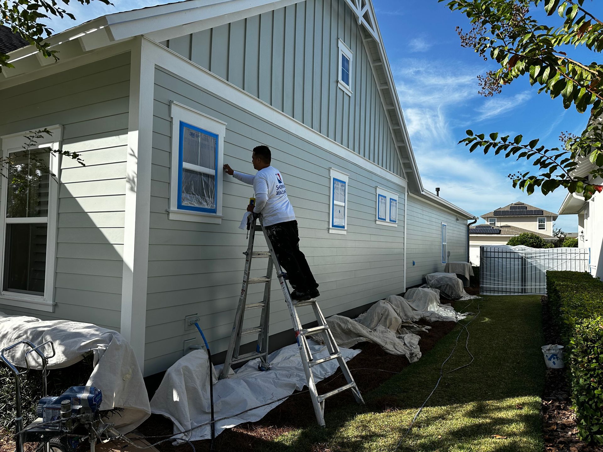 A man is standing on a ladder painting the side of a house.