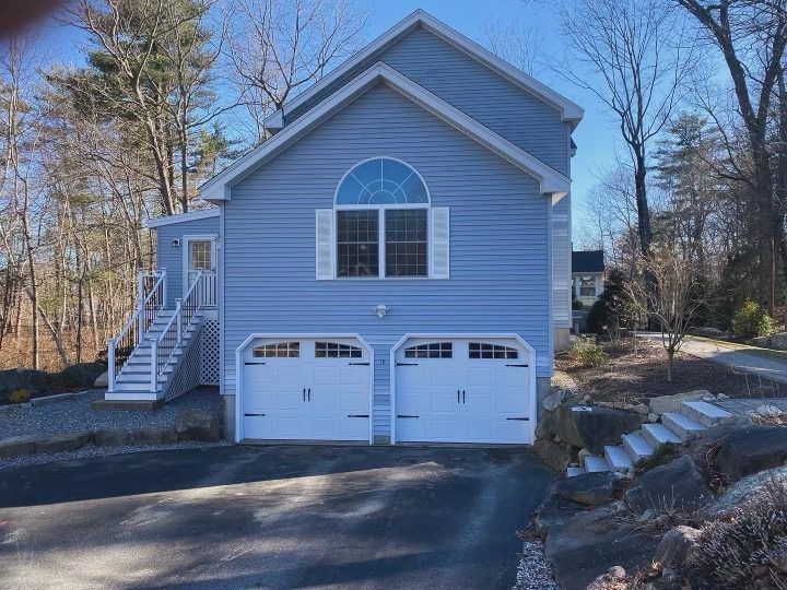 A two-story light blue house with a two-car garage, stairs to the entrance, and trees in the background under a blue sky.