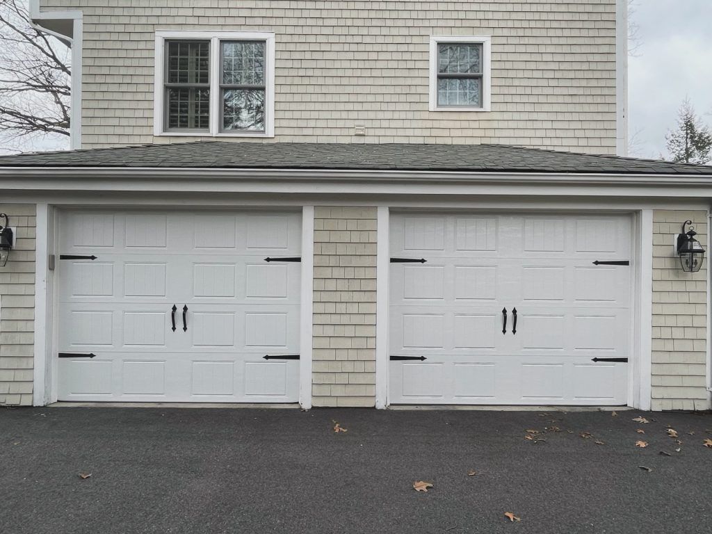 Two white, paneled residential garage doors with black decorative hardware on a house with beige siding.