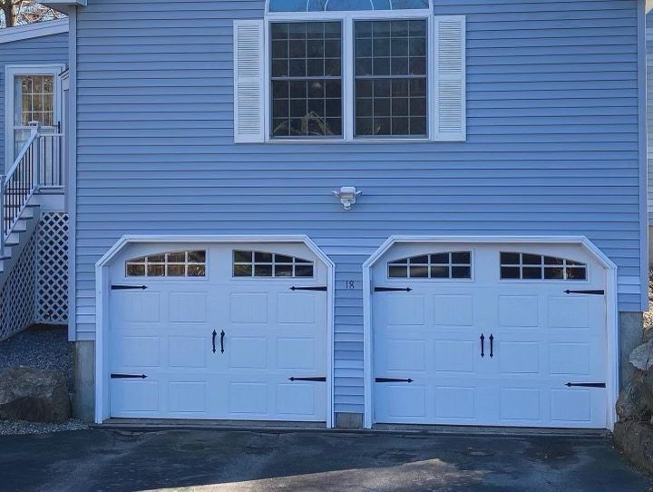 A two-car white garage door with decorative black hinges and windows, set into the light blue siding of a home.