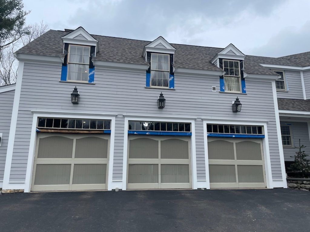 A light gray house with three garage doors, three dormer windows, and exterior lanterns on an asphalt driveway.