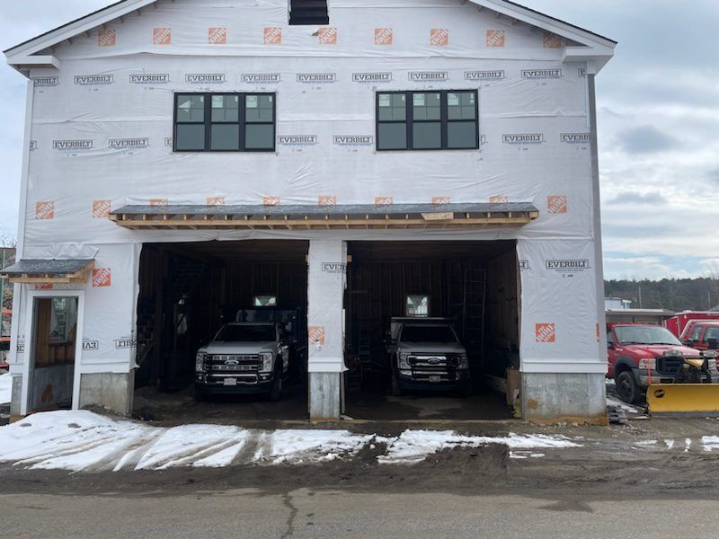 A two-story building under construction with two garage bays, each containing a pickup truck, under a cloudy sky.