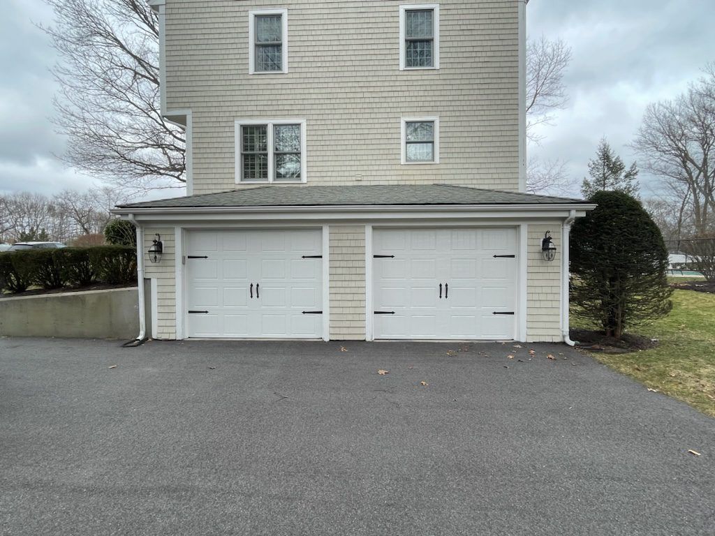 A two-story light-colored house with a two-car garage at street level, situated on an asphalt driveway under cloudy skies.