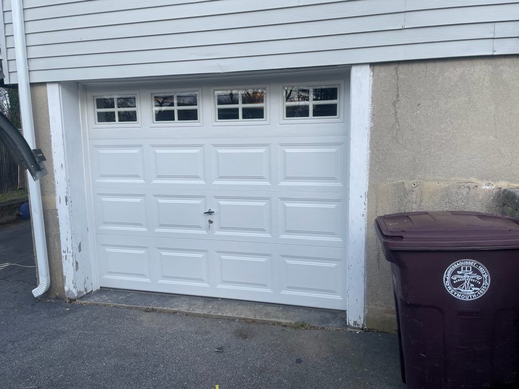A white paneled garage door with top windows, set in a house with light siding and a dark trash bin to the right.