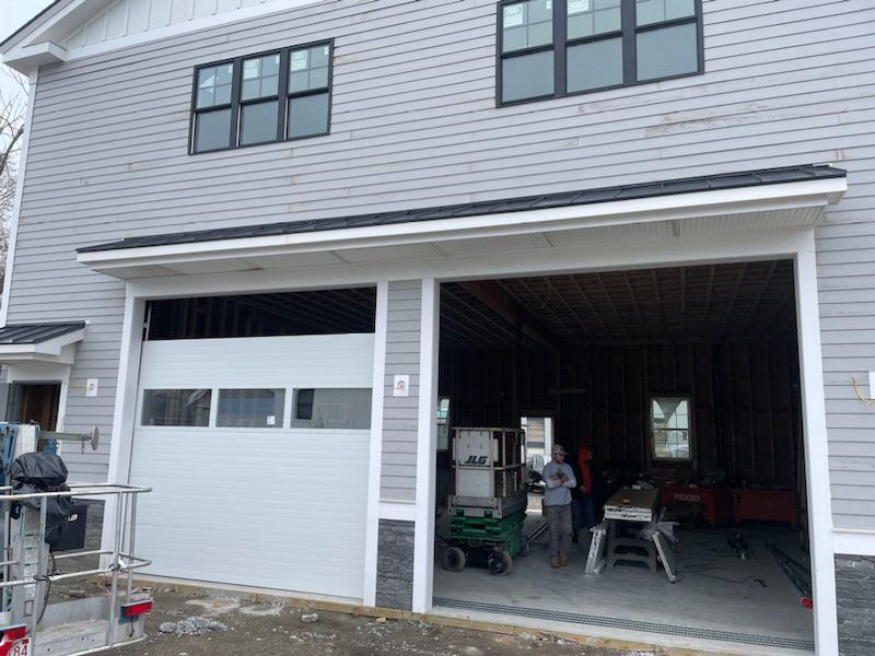 A two-story gray building under construction with one installed garage door, one open bay, and people inside.