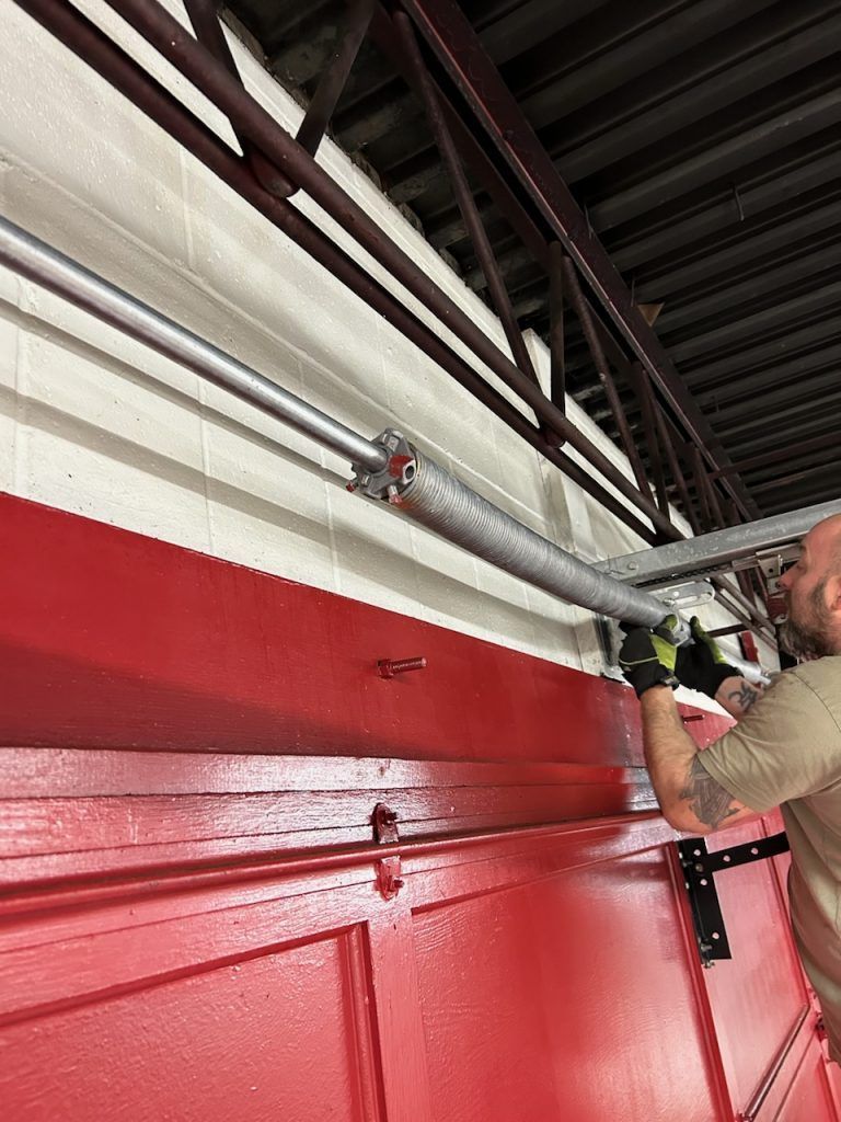 A worker in gloves adjusts a metal spring on a garage door torsion bar mounted above a red door.