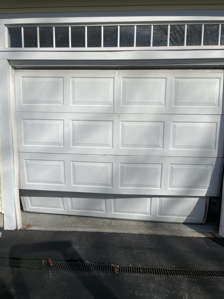 A white garage door is partially open, with the bottom panel misaligned and detached from the track.