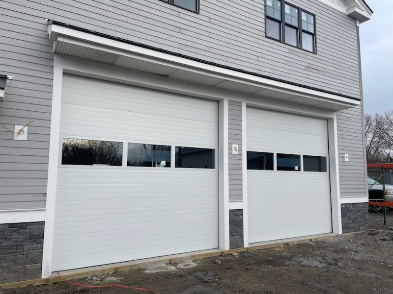 Exterior of a building featuring two modern white garage doors with rectangular windows, gray siding, and stone accents.