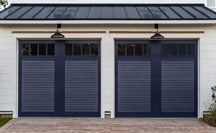 Two dark navy garage doors with horizontal slats and top glass panels, set in a white wall with metal lanterns.