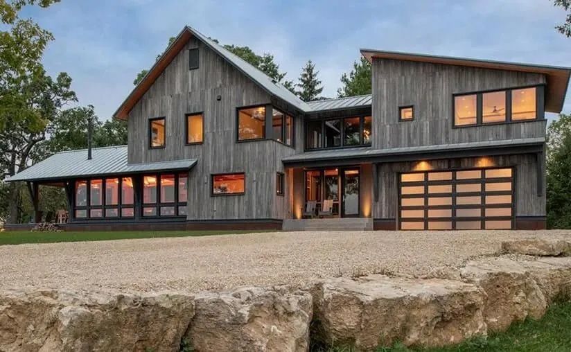 A modern, two-story house with vertical grey wood siding, a metal roof, and large windows, situated above a stone wall.