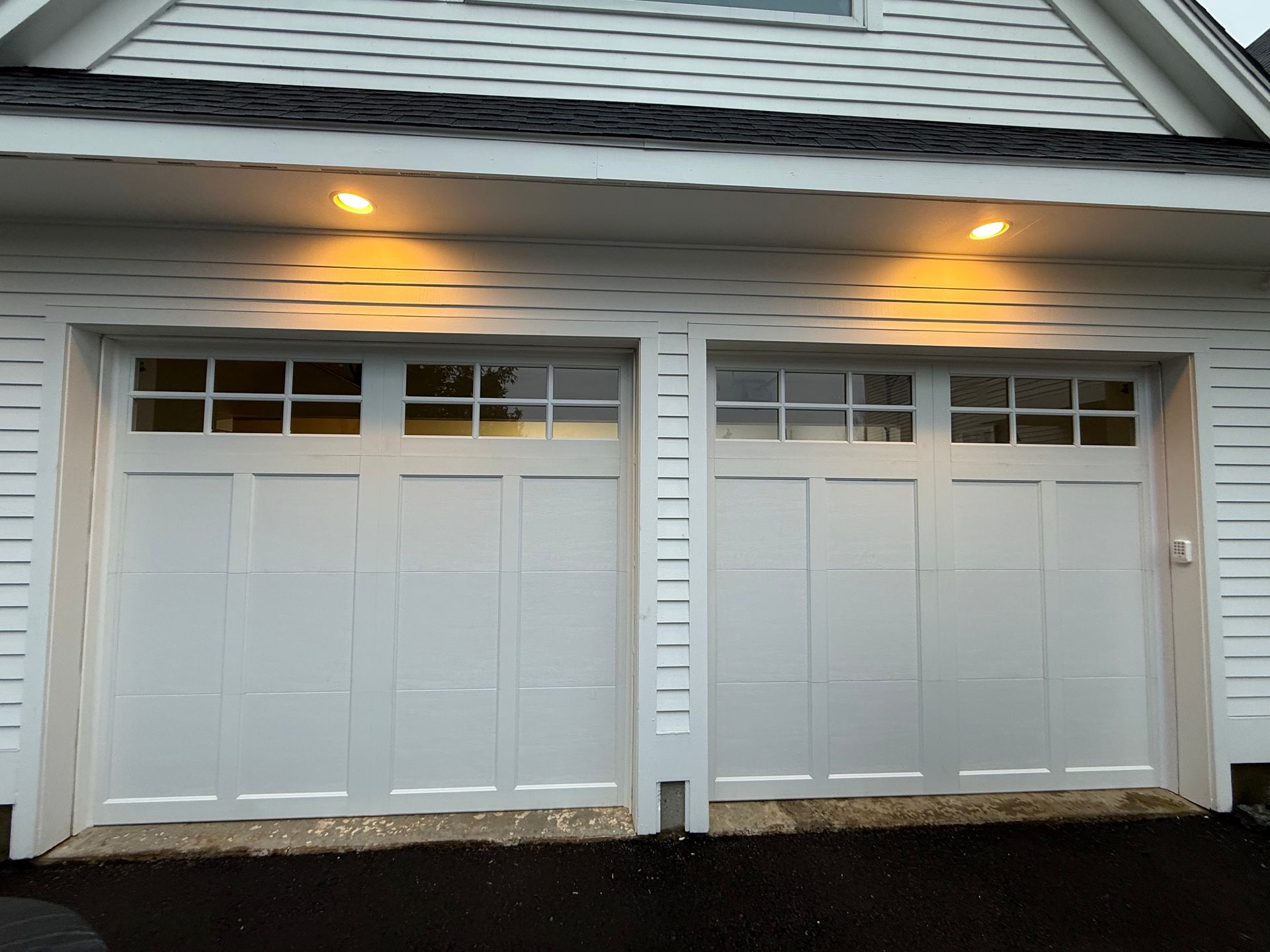 Two white, four-panel carriage-style garage doors with windows, illuminated by outdoor lights above.