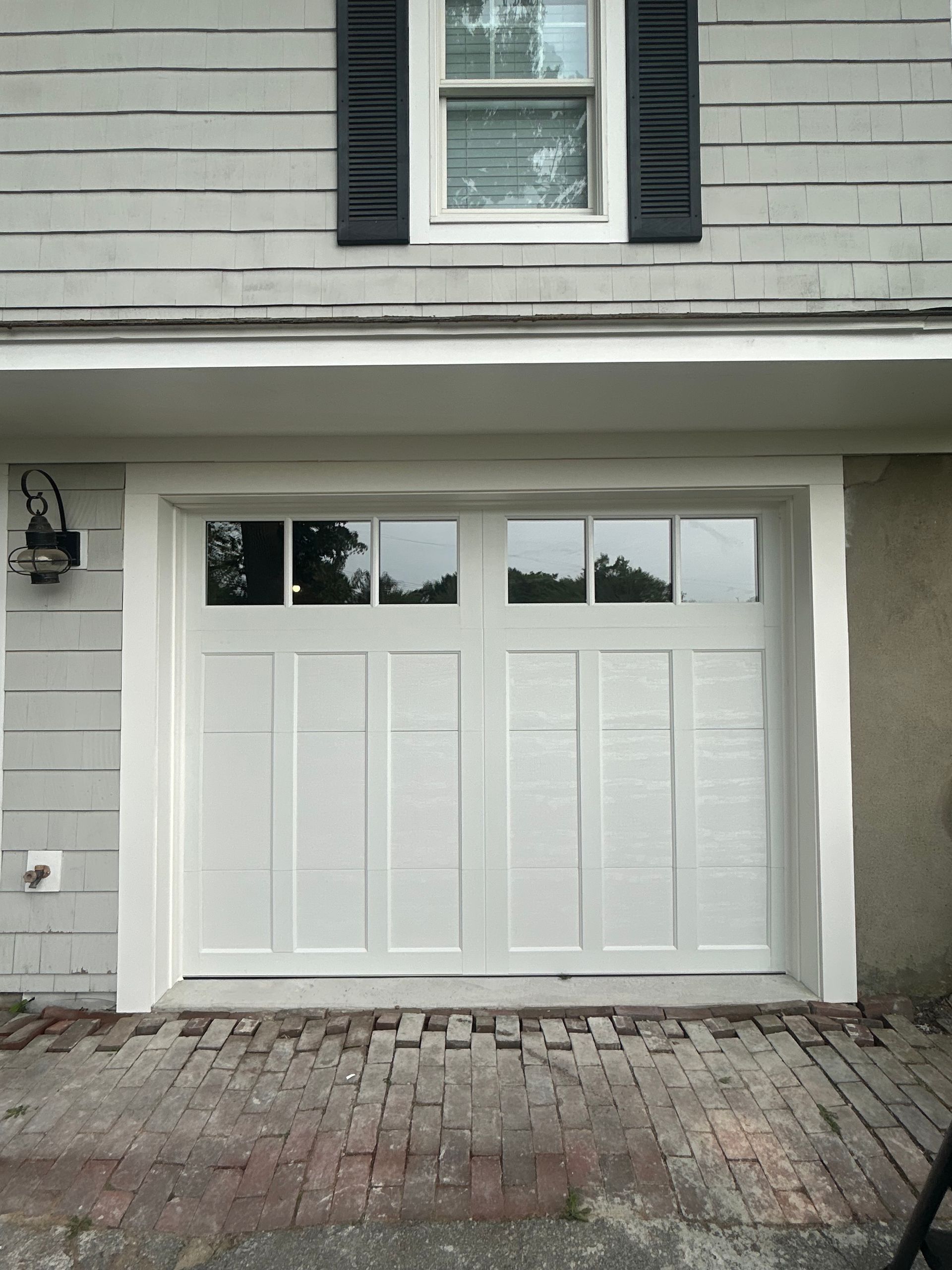White garage door on a gray house with a brick driveway and a window above.