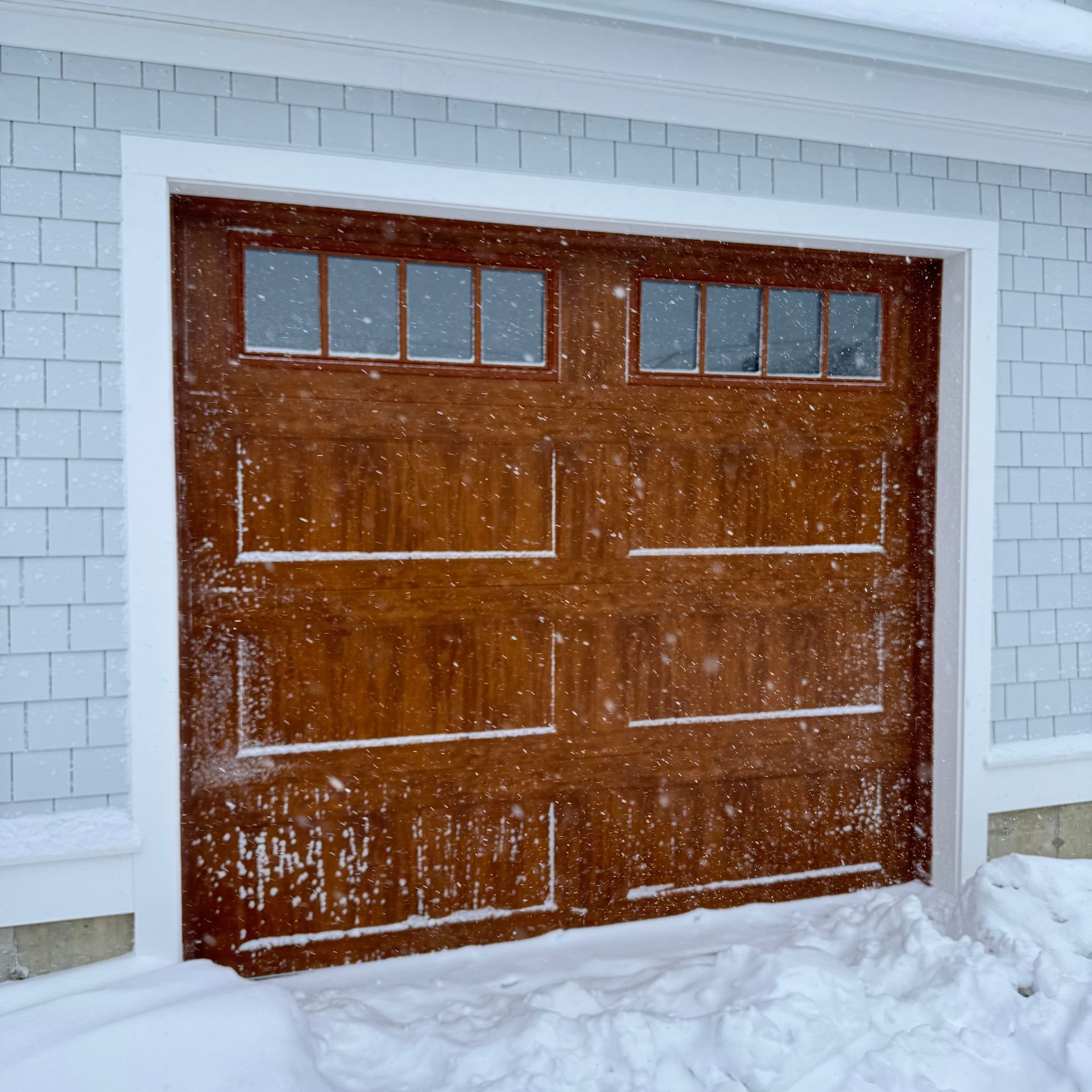 A wood-tone garage door with two window sections, framed in white trim against light-blue siding, during a light snowfall.
