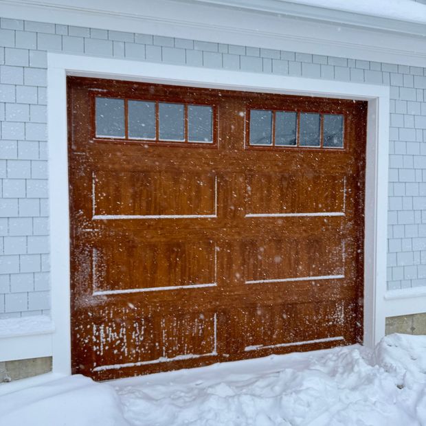 A wood-tone garage door with two window sections, framed in white trim against light-blue siding, during a light snowfall.