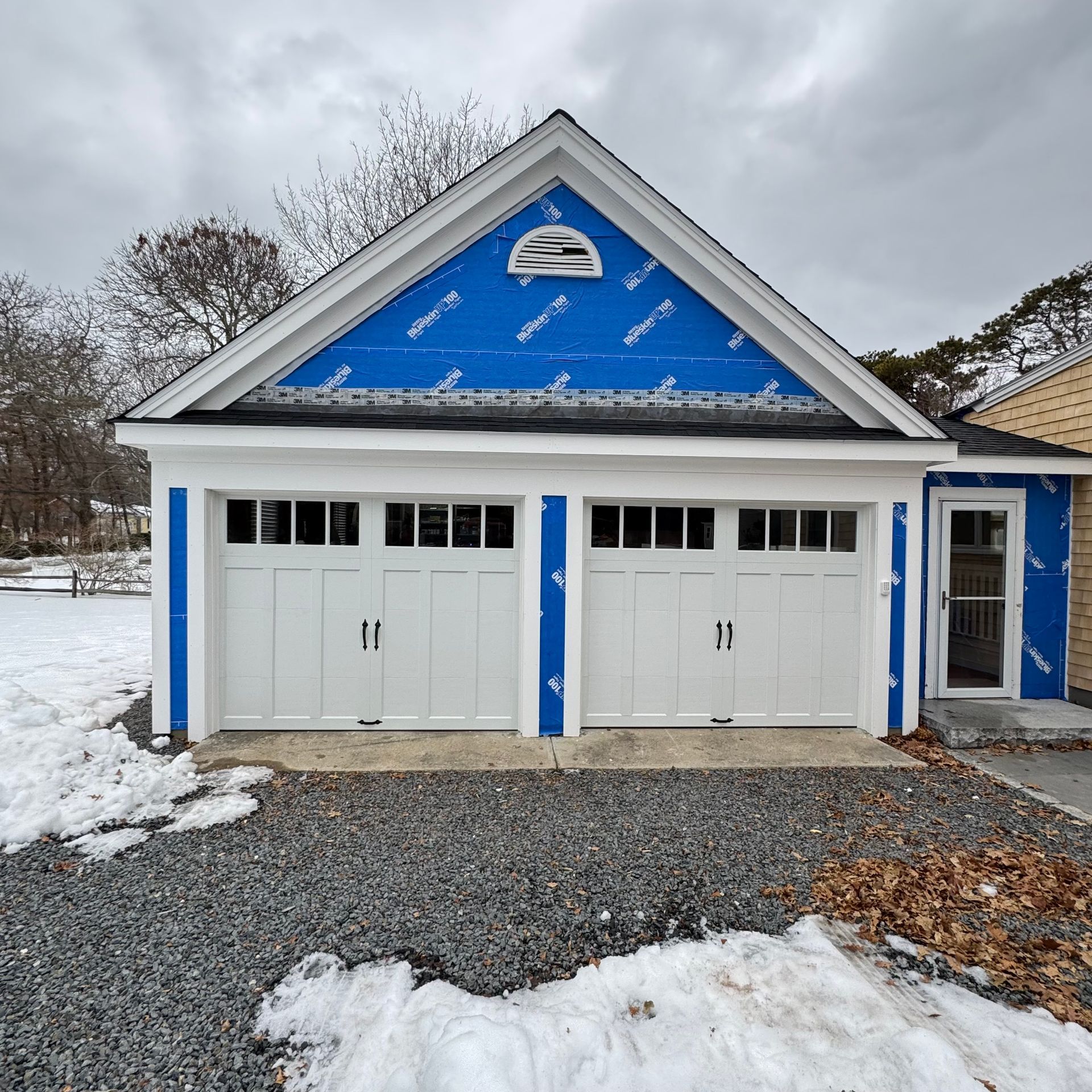 A two-car garage under construction with white doors, blue weather-resistant house wrap on the gable, and a gravel driveway.