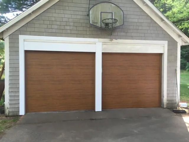 A gray shingled garage with two wood-look doors and a basketball hoop mounted above them.