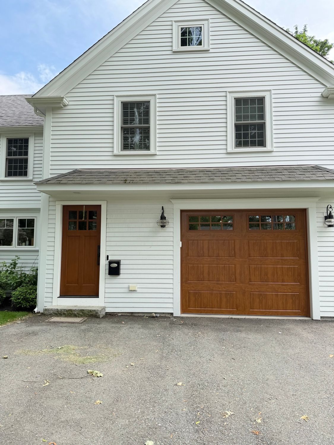 White two-story house exterior with a wooden front door, garage door, and gravel driveway