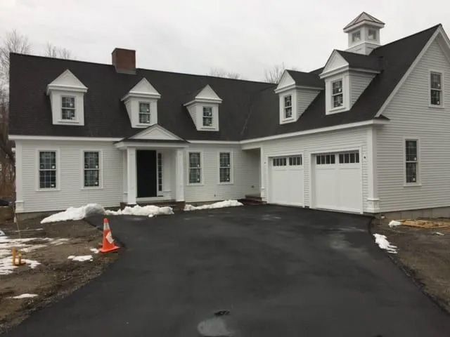 A newly built, two-story white house with a dark roof, dormer windows, and a two-car garage, seen from a paved driveway.