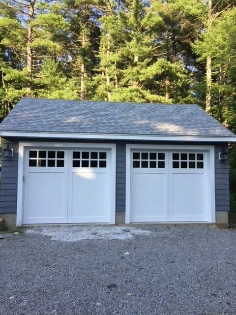 A detached two-car garage with white carriage-style doors and grey siding, situated in a wooded area with a gravel driveway.
