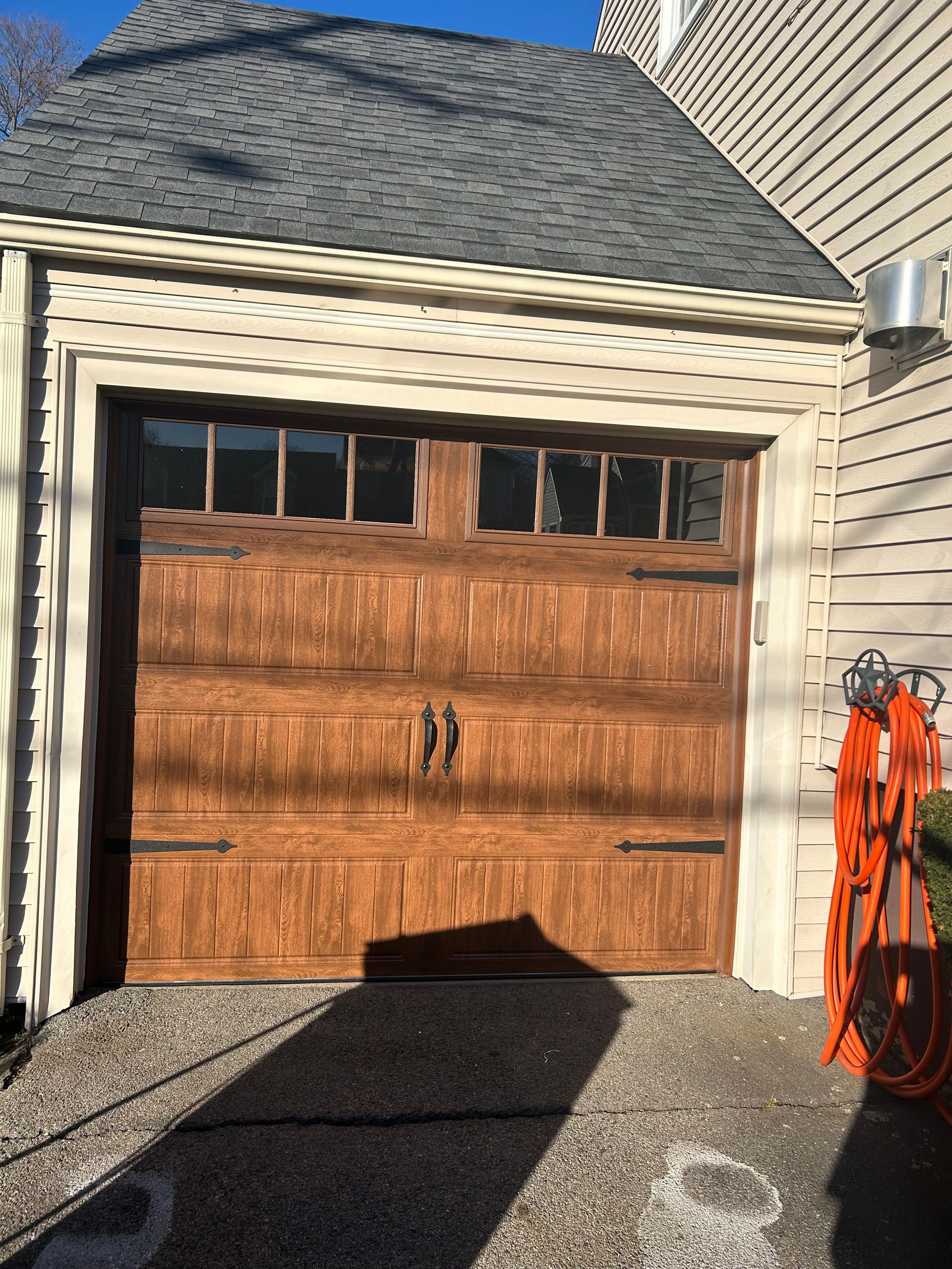A wooden carriage-style garage door with glass panes at the top, mounted on a house with beige siding and a shingled roof.