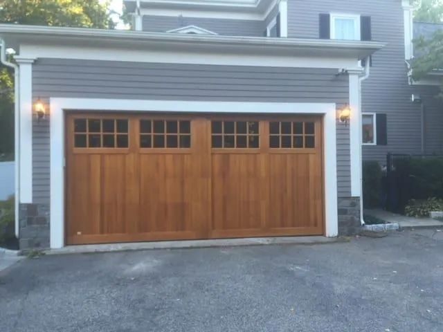 A two-car wooden garage door with windows on a house with gray siding and stone accents.