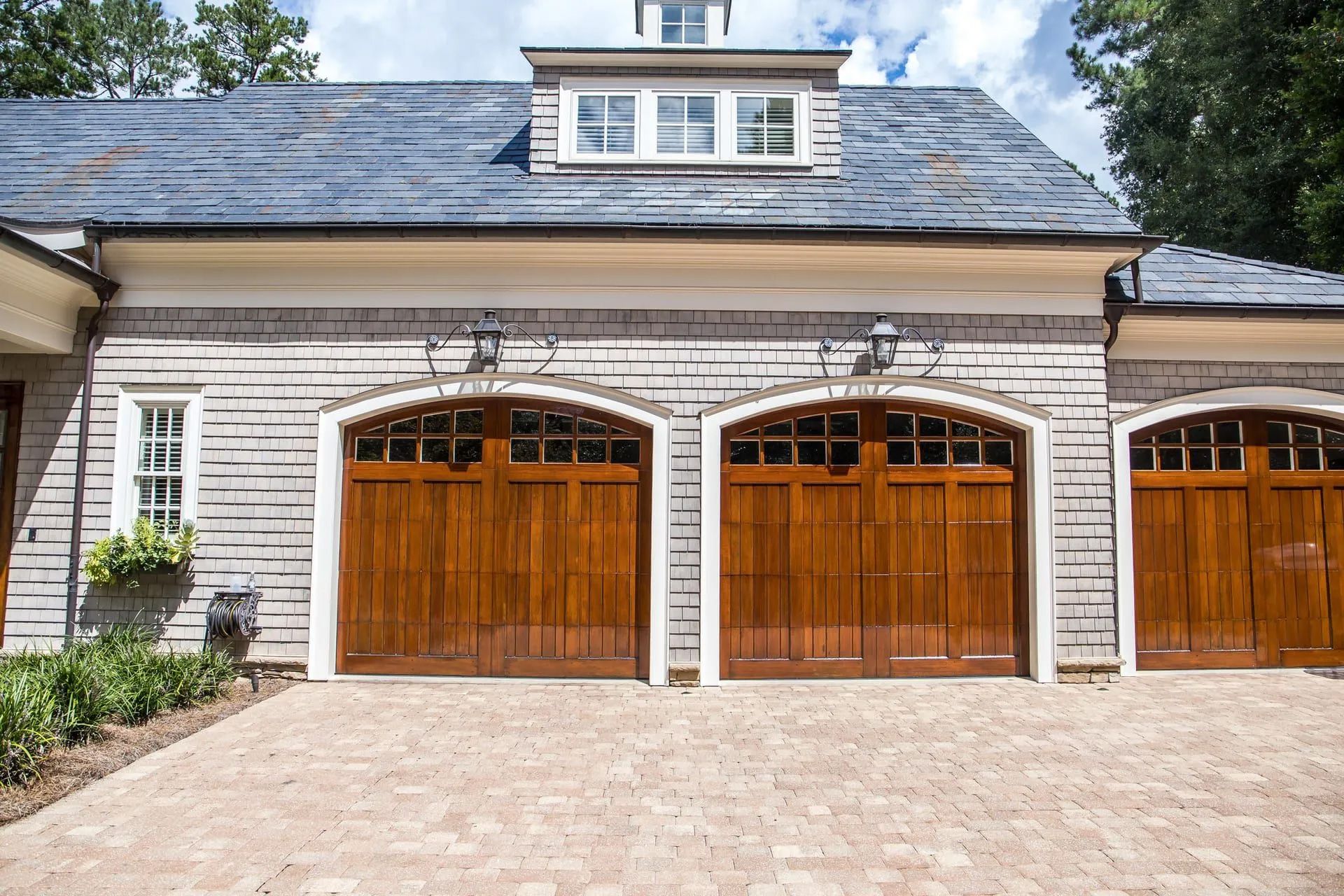 A stone house with three polished wooden garage doors, a dormer window, and a light-colored paved driveway.