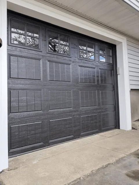 A dark gray, paneled garage door with top-row windows set in a white frame against a light-colored house siding.
