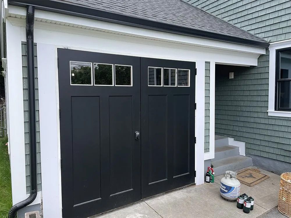 Black double garage doors with windows, set in a white frame against green shingled siding with concrete steps nearby.