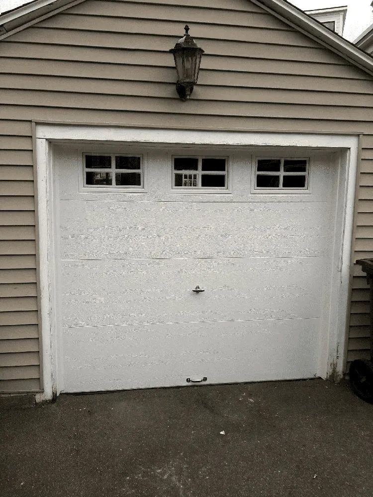A white garage door with three small, square windows at the top, mounted beneath a wall-mounted outdoor lamp.