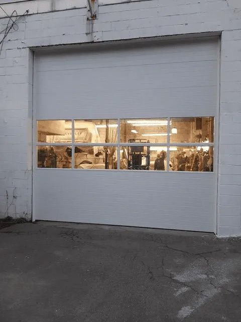 A white sectional garage door with a row of clear glass windows at eye level, showing a brightly lit indoor workshop.