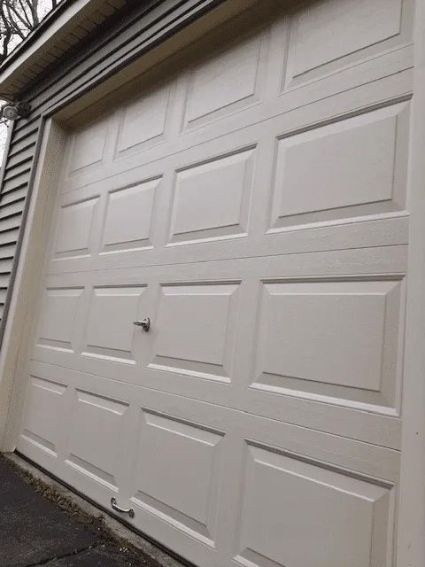 A beige residential garage door with a raised panel design, a central handle, and a bottom pull handle.