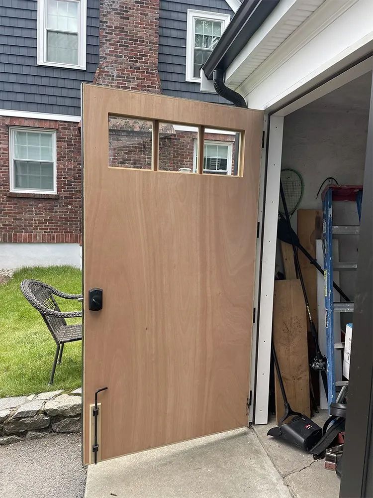 A partially open plywood door with three small, horizontal windows installed in a garage frame beside a brick house.