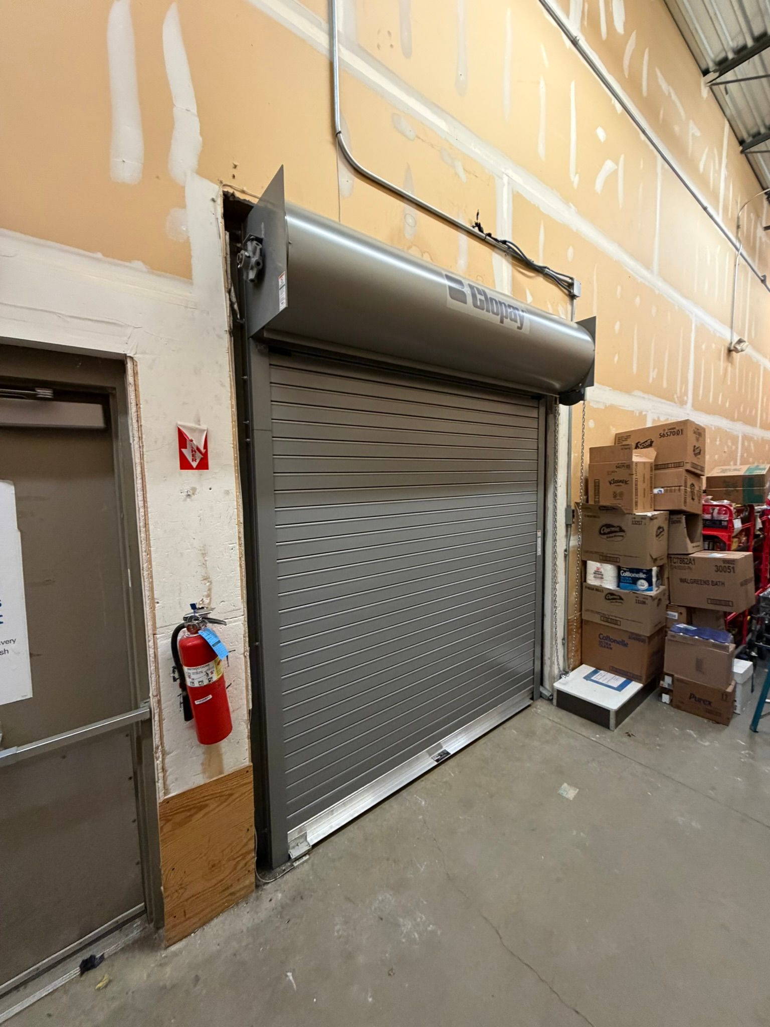 Closed metal roll-up door beside stacked boxes in a warehouse corridor, with a fire extinguisher on the wall.