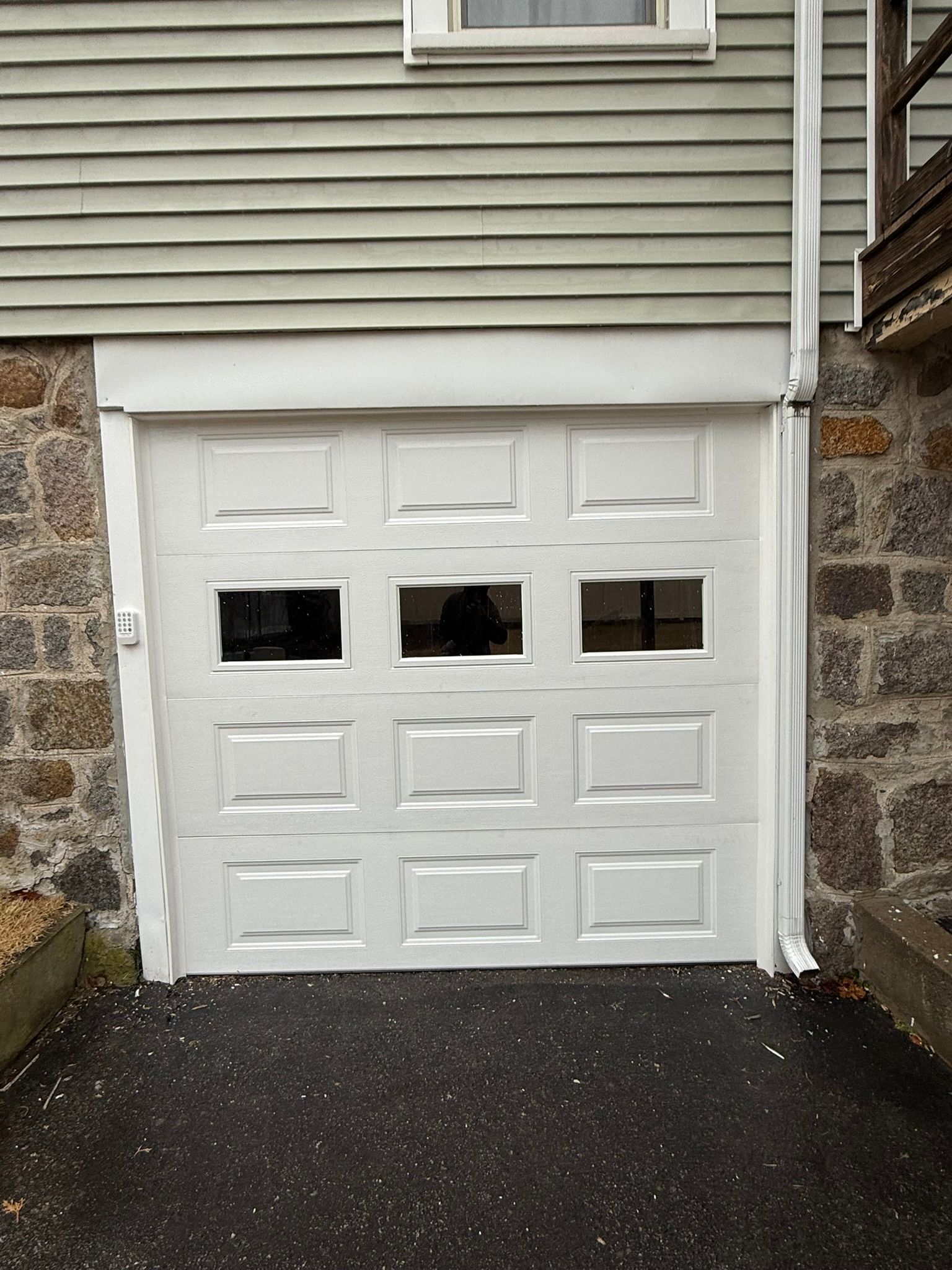 White garage door with three small windows under gray siding and stone walls, on a paved driveway