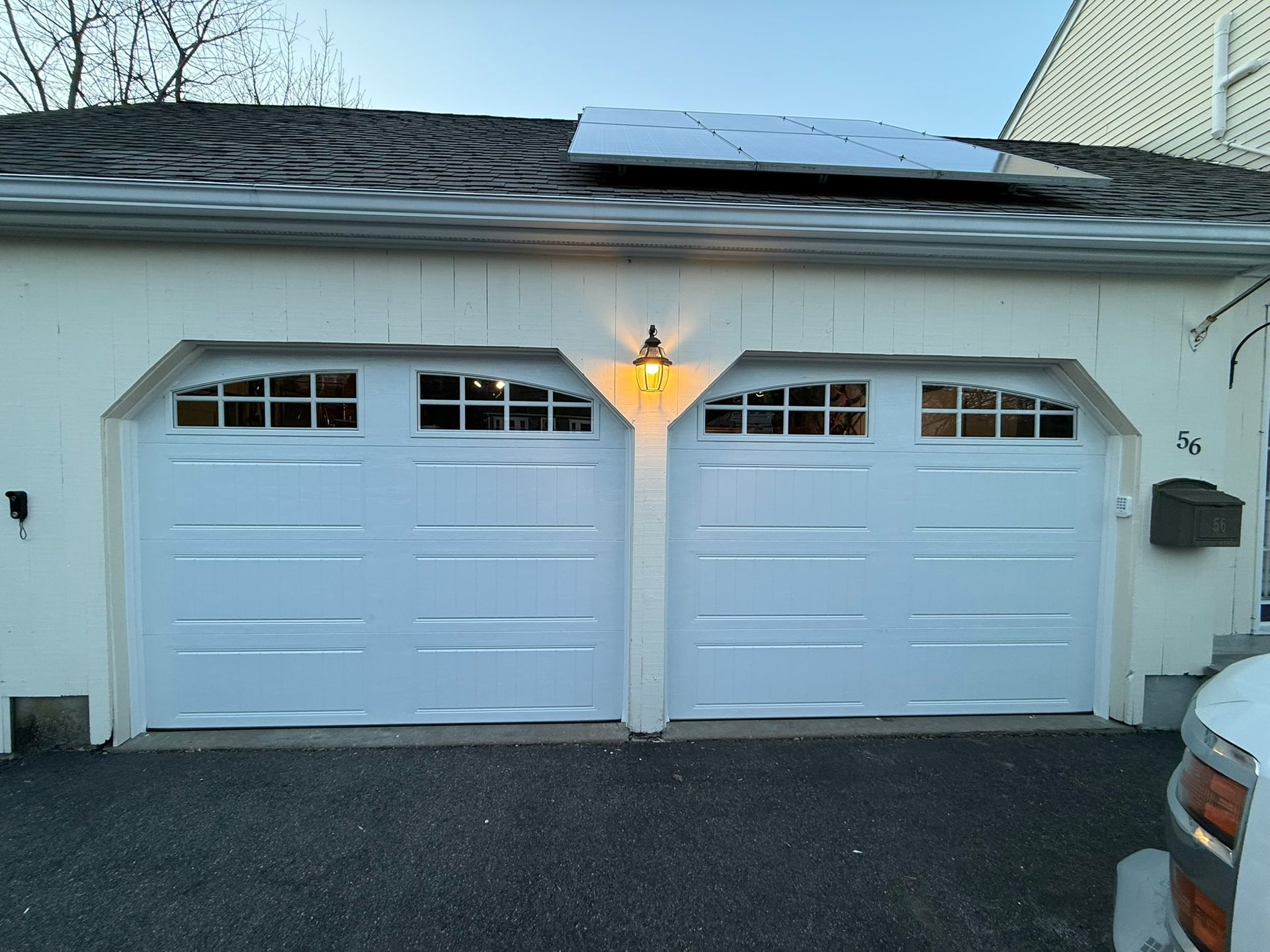 Two white garage doors with arched windows under a lit porch lamp at dusk