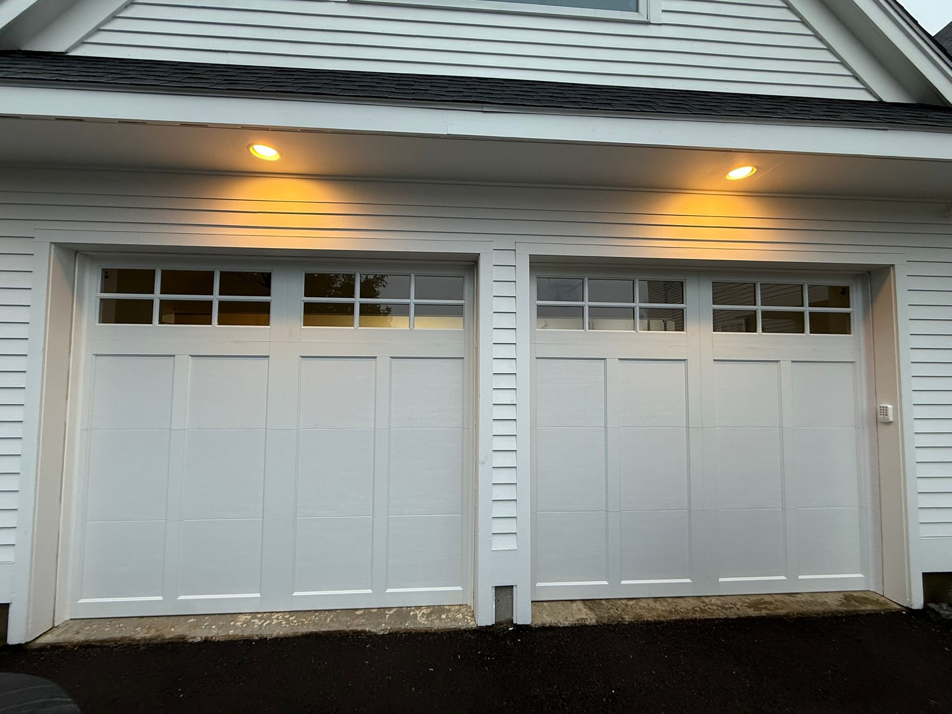 Two white garage doors on a house exterior with lit porch lights at dusk