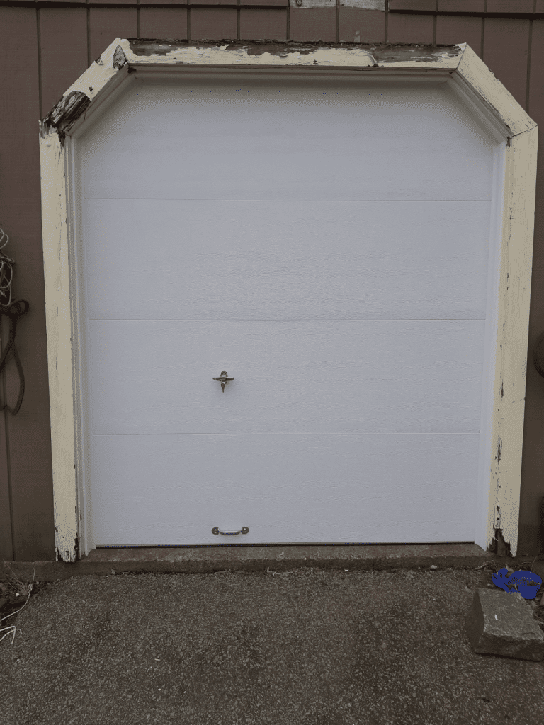 A white, rectangular garage door with angled top corners, set within a weathered, light-colored wooden frame.