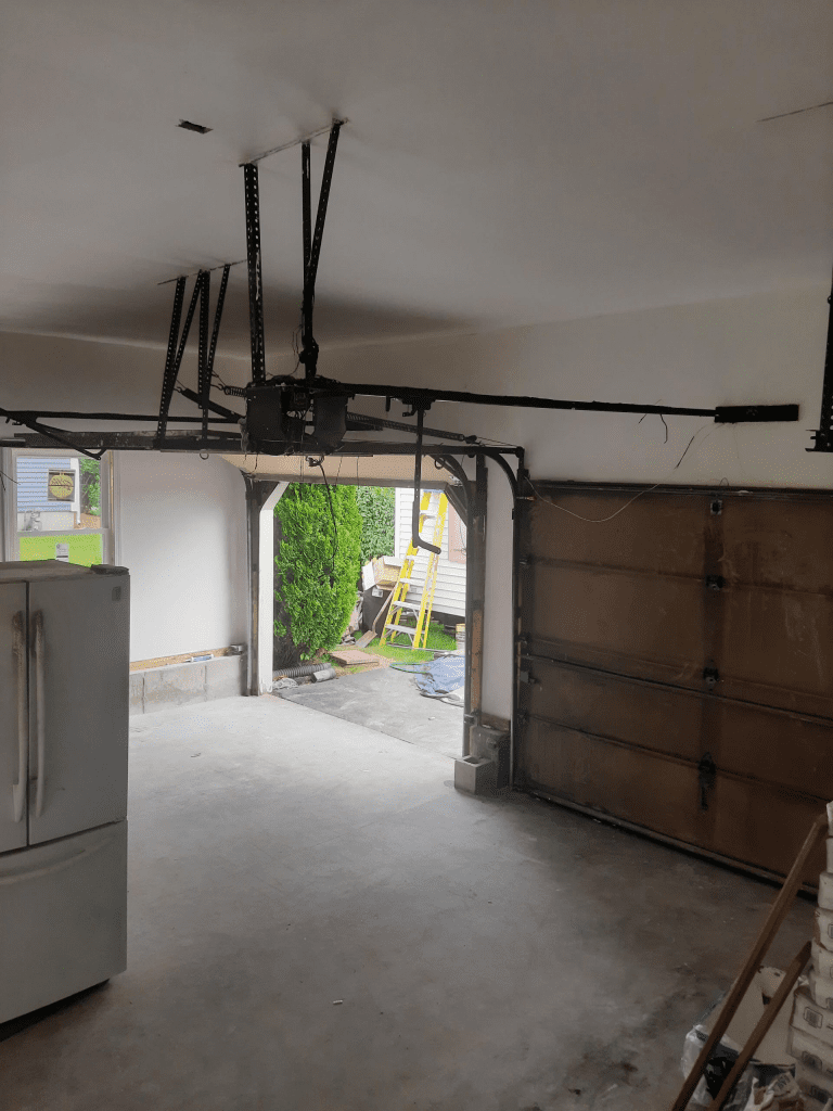 A view inside a garage with a ceiling-mounted door opener, a white refrigerator on the left, and an open door to a yard.