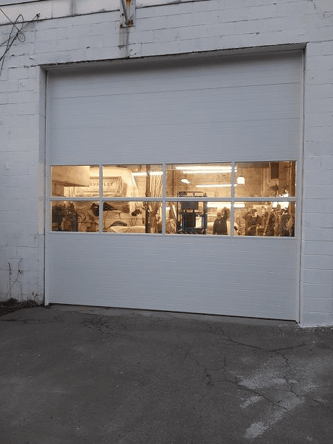 A white commercial garage door with a central row of rectangular glass windows looking into a lit workshop interior.