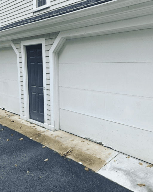 A dark gray door set into the wall between two white garage doors, located at the edge of a paved driveway.