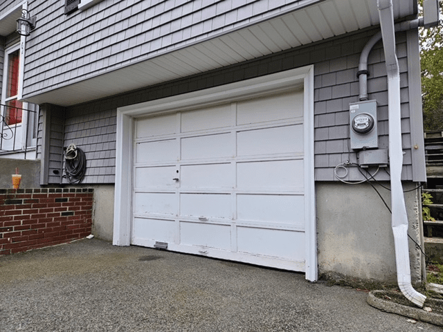 White garage door on a grey-shingled house with a brick planter and utility meter on the exterior.