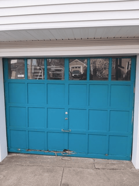A blue, multi-panel garage door with a row of windows at the top, showing significant wood rot at the bottom center.