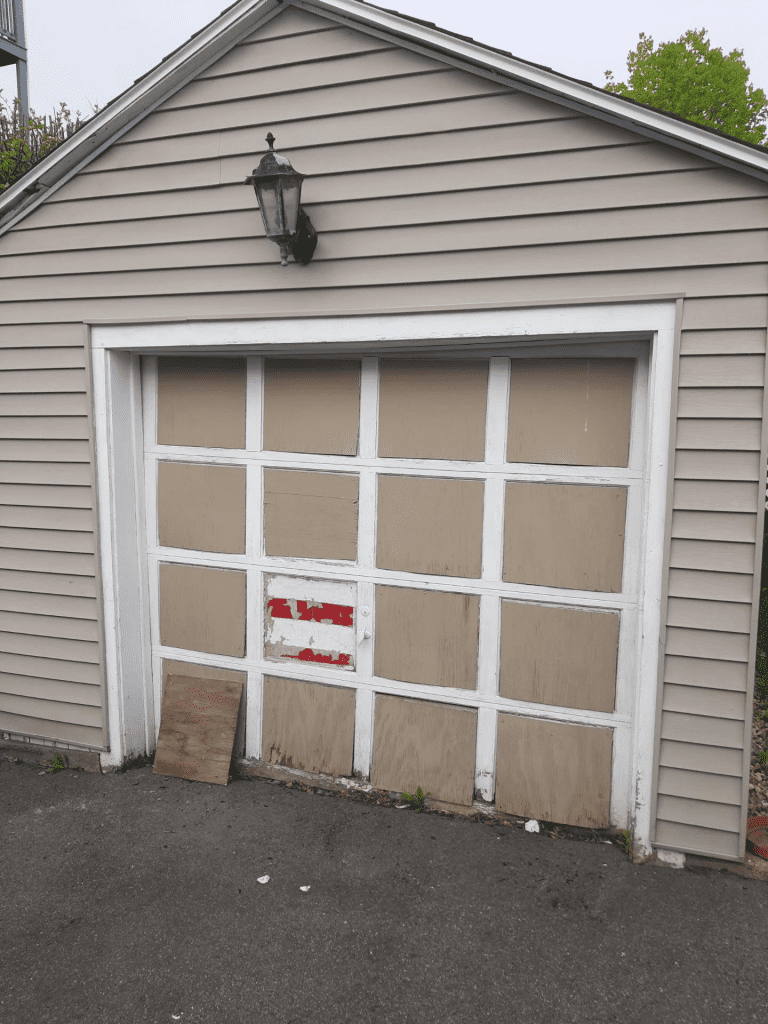 A detached garage with beige siding, a wall-mounted lamp, and a white-framed door with patchy brown and red panels.