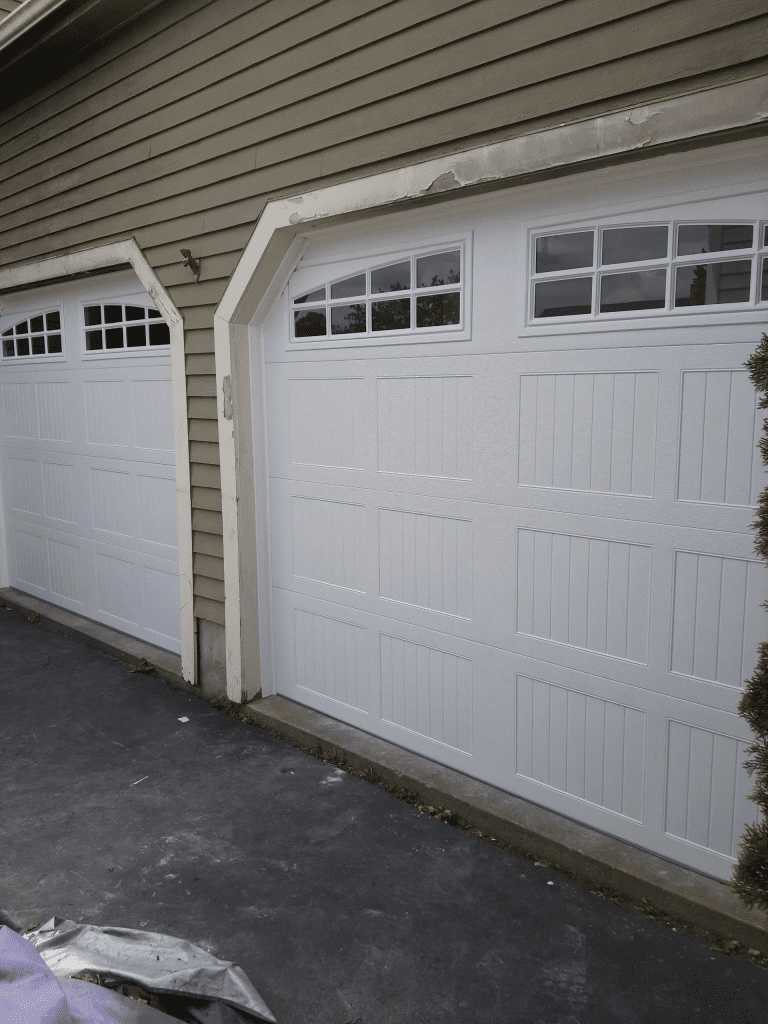 Two white garage doors with rectangular windows along the top, set into a house with tan siding and a dark paved driveway.
