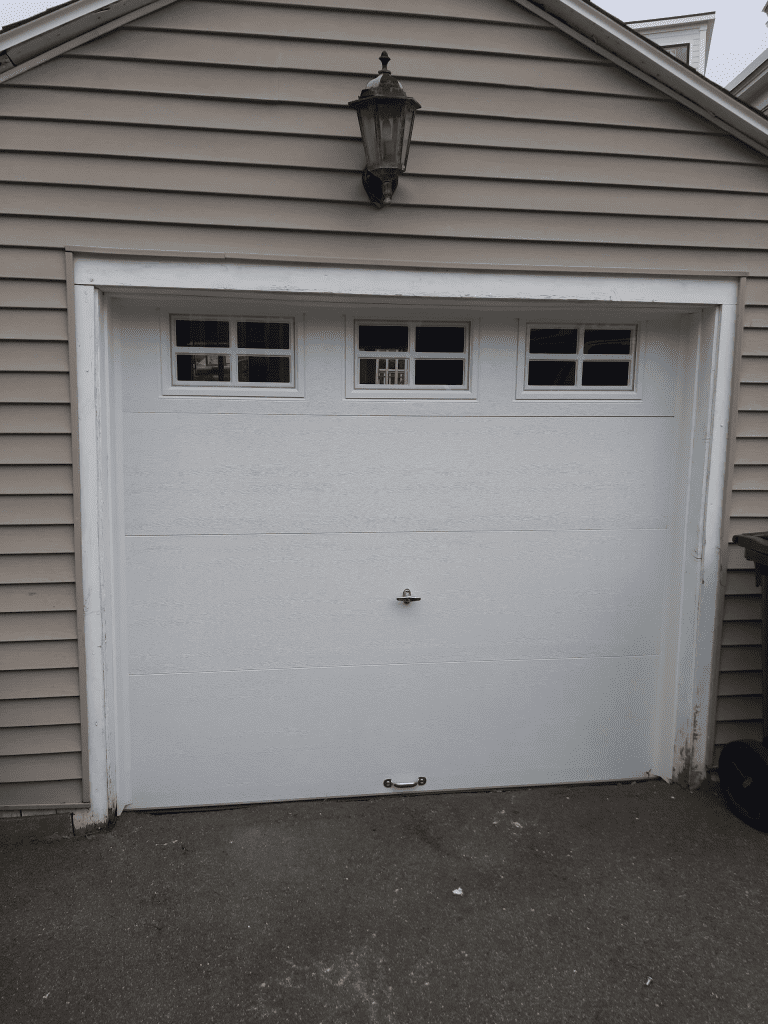 A white single-car garage door with three small windows and a metal handle, set in a house with light tan siding.