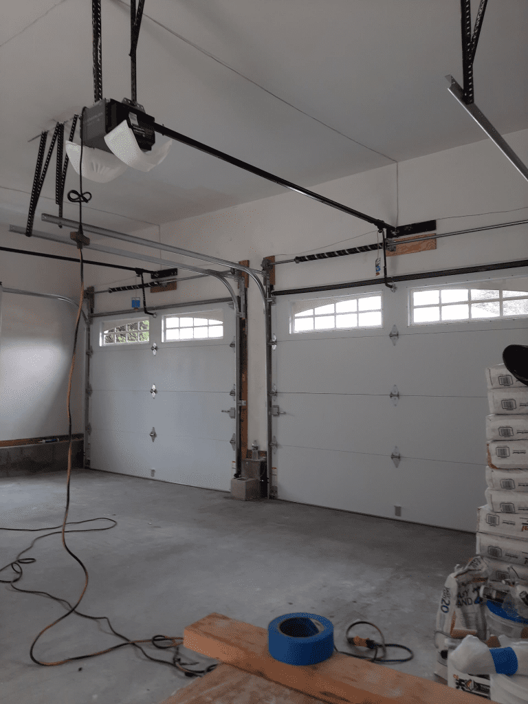 An interior view of a garage with two white sectional garage doors, overhead openers, and construction materials.